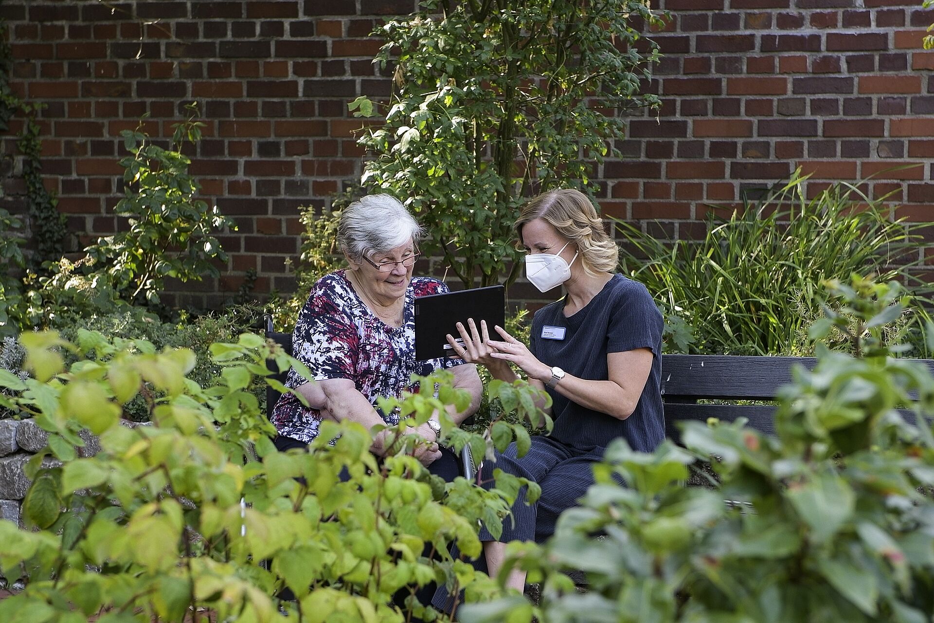 Ein Mitarbeiterin des sozialen Dienst sitzt mit einer Bewohnerin im Garten und hält ein Tablet